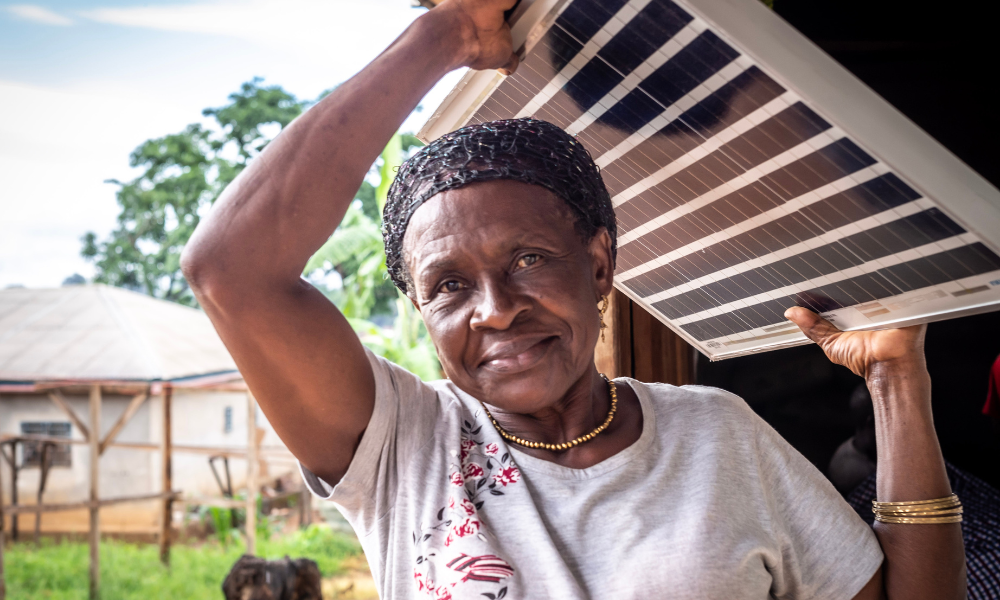 Niembain Charlotte, a 60 year old grandmother and widow from Cameroon stands smiling holding a solar panel above her head at an angle. She was trained as a solar engineer by Barefoot College International and has installed over 400 solar home lighting systems in rural and remote communities.