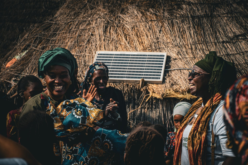 A group of rural women in Senegal stand in front of a house that has been electrified with solar energy. They are celebrating and dancing.